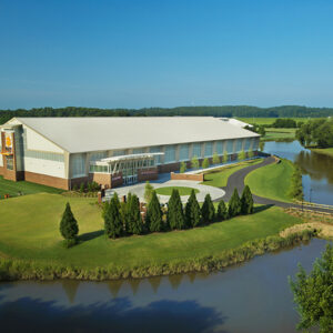 Aerial view of a large building surrounded by trees, a circular driveway, and a river on a sunny day.