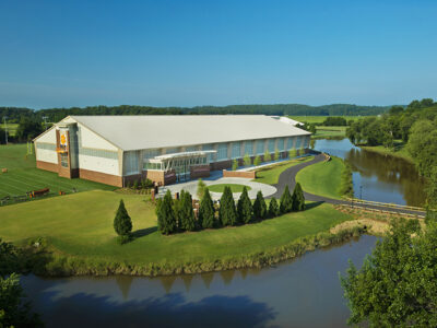 Aerial view of a large building surrounded by trees, a circular driveway, and a river on a sunny day.