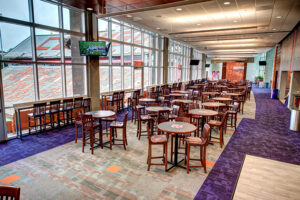 Spacious room with numerous round tables and chairs, large windows, television, purple carpet, and a view of a stadium seating area.