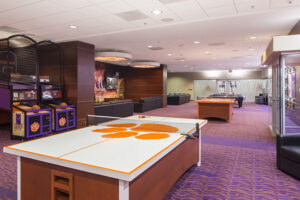 A recreation room with ping pong tables, arcade basketball games, and couches on a purple carpeted floor.