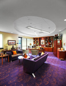 Modern office with bookshelves, desk, two purple sofas, and a table on a patterned carpeted floor, under a circular ceiling design.