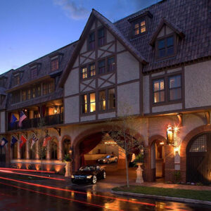 Exterior of a Tudor-style building at dusk with lit windows, balconies, and several flags. A car is parked in front.