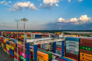 Stacks of colorful shipping containers with cranes at a port under a partly cloudy sky.