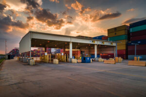 Shipping containers stacked near a port building under a cloudy sunset sky.