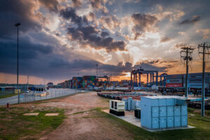 Container terminal at sunset with stacks of shipping containers, cranes, utility boxes, and dramatic clouds in the sky.