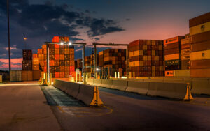 Shipping containers stacked at a port during dusk, with illuminated streetlights and a cloudy sky in the background.