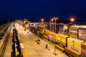 Aerial view of a well-lit shipping container yard at night with stacked containers and handling vehicles.