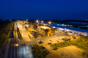 Aerial view of a container terminal at night, with stacked shipping containers, cranes, and illuminated infrastructure.