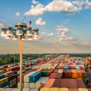 View of a large shipping container yard with rows of colorful containers under a partly cloudy sky and a tall light pole.