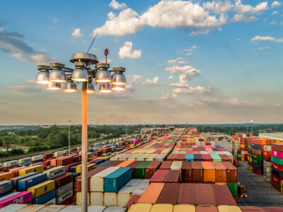 View of a large shipping container yard with rows of colorful containers under a partly cloudy sky and a tall light pole.