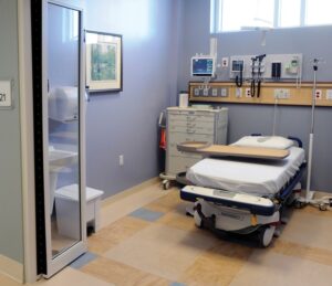 Hospital room with a bed, medical equipment, a wall-mounted monitor, and a window. A supply cabinet is visible on the left.