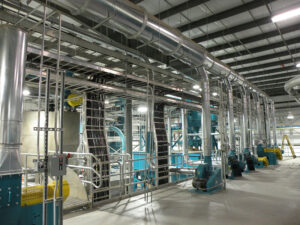 Industrial facility interior with large pipes, machinery, and metal walkways under a high ceiling.