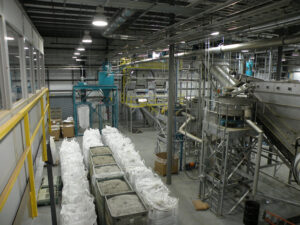 Interior of an industrial recycling plant with bags of materials, processing machinery, and overhead metal structures.