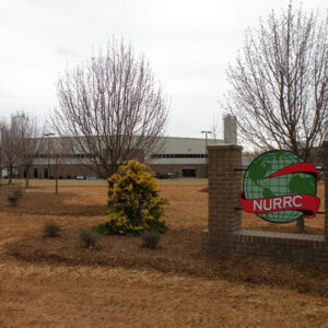 A large industrial building with a "NURRC" sign in front, surrounded by trees and a grassy area.