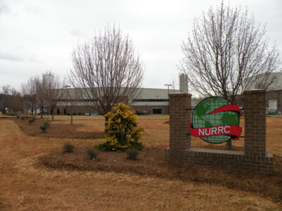 A large industrial building with a "NURRC" sign in front, surrounded by trees and a grassy area.