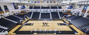 An empty indoor basketball arena with "Wofford" on the court, surrounded by tiered seating and large screens.