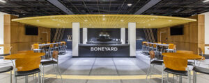 Interior of a modern cafeteria with "Boneyard" signage, featuring round tables, chairs, and a central counter under a yellow ceiling.