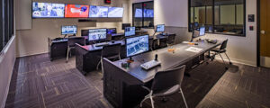 A control room with multiple monitors on desks displaying video feeds and equipment, surrounded by large windows and screens.