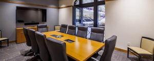 A conference room with a rectangular wooden table, surrounded by ten black chairs, and a wall-mounted TV screen.