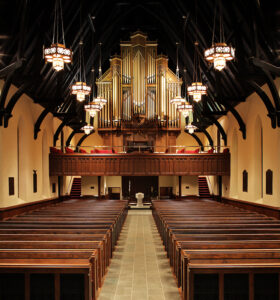 Interior of a church with wooden pews, an ornate pipe organ, chandeliers, and a central aisle leading to the altar.