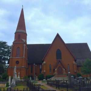 A brick church with a tall steeple stands amidst an old graveyard under a cloudy sky.