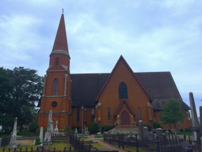 A brick church with a tall steeple stands amidst an old graveyard under a cloudy sky.