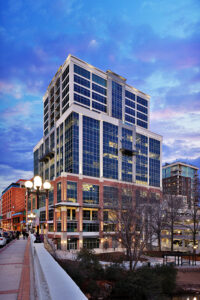 A modern high-rise building with glass and brick exterior under a cloudy sky, located next to a street with parked cars.