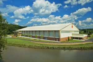 Large sports facility with a white roof, brick exterior, and emblem on the facade, set by a waterway under a blue sky with clouds.