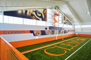 Indoor football practice facility with "Clemson" on the walls and field, featuring images of tiger eyes and team logos.