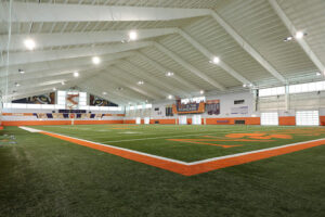 Indoor football training facility with artificial turf, white ceiling, and bright lighting. Orange and white decor accents the space.