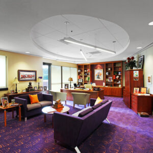 Modern office with purple carpet, two sofas, a desk, bookshelves, and framed photos on walls in a well-lit room.