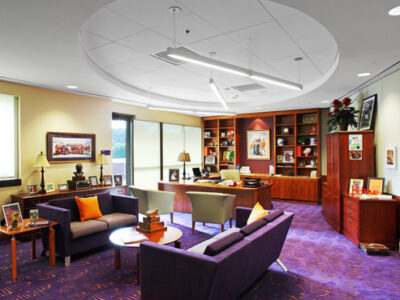 Modern office with purple carpet, two sofas, a desk, bookshelves, and framed photos on walls in a well-lit room.