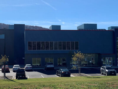 A modern building with large windows and a parking lot with several parked cars under a clear blue sky.