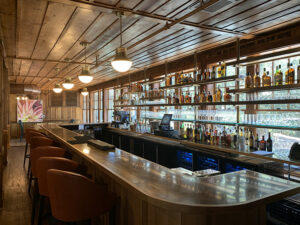 A wooden bar with shelves of bottles, leather stools, pendant lights, and a painting on the wall.