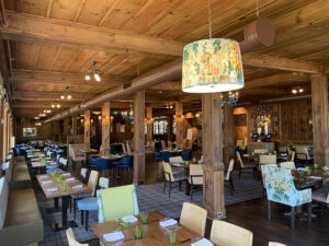 Spacious restaurant interior with wooden ceiling, tables set with green glasses, and patterned chairs under decorative lighting.