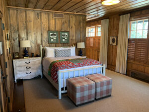 A rustic bedroom with wood-paneled walls features a bed, plaid ottomans, a dresser, lamps, and shuttered windows.