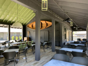 Outdoor dining area with empty tables and chairs, wooden canoe hung on a wall, and a green ceiling under a covered patio.