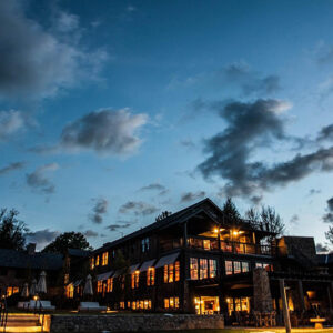 A large building with lit windows stands under a cloudy evening sky, surrounded by trees and outdoor seating.