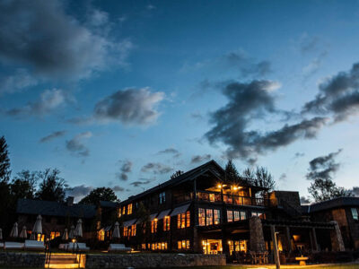 A large building with lit windows stands under a cloudy evening sky, surrounded by trees and outdoor seating.