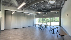 Empty classroom with foldable tables and chairs, large windows, and gray partition walls under ceiling lights.