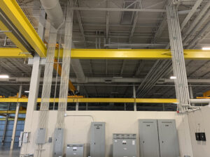 Industrial interior with yellow beams, hanging cables, and electrical panels against a white wall and high ceiling.