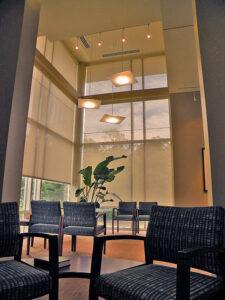 Modern waiting room with high ceilings, large windows, hanging lights, and arranged seating, featuring a large potted plant.