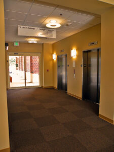 Hospital corridor with three elevators on the right and a glass entrance door at the end, signs hanging from the ceiling.