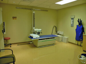 Empty hospital X-ray room with a table, protective aprons on the wall, and an X-ray machine.