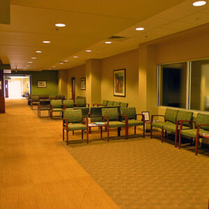 A spacious waiting room with rows of empty chairs, beige walls, framed art, and overhead lighting.