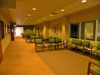 A spacious waiting room with rows of empty chairs, beige walls, framed art, and overhead lighting.