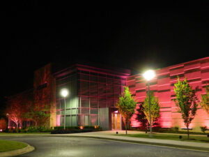 Modern building illuminated with pink and white lights at night, surrounded by trees and a streetlamp in the foreground.