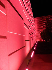 Exterior wall with geometric patterns illuminated by pink lights at night, with a pathway and shrubs alongside.