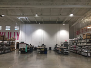 A warehouse interior with shelves of boxes, workstations, and overhead lighting. The floor is polished concrete.