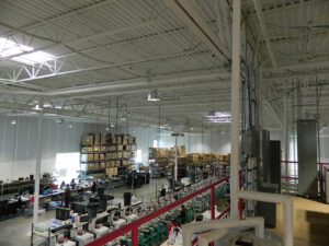 Interior of a large warehouse with stacked boxes, workstations, and high ceilings with hanging lights.